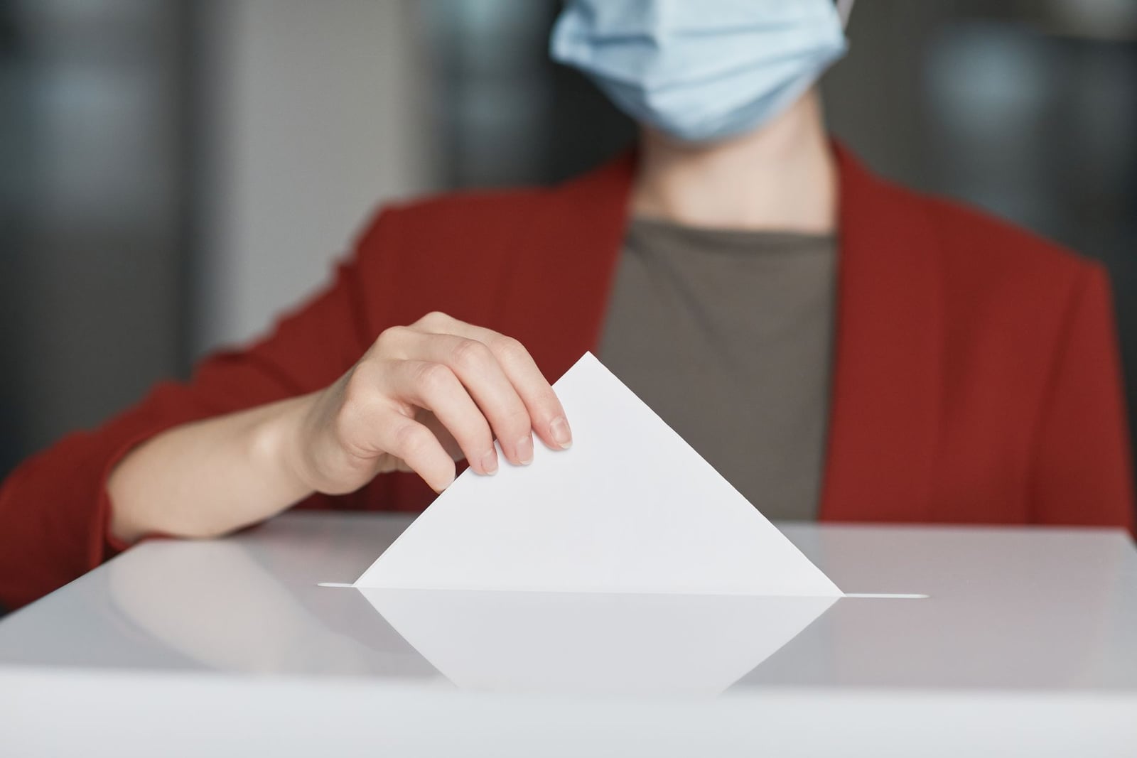 Persona con mascarilla depositando un voto en una urna. Enfatiza el deber cívico.
