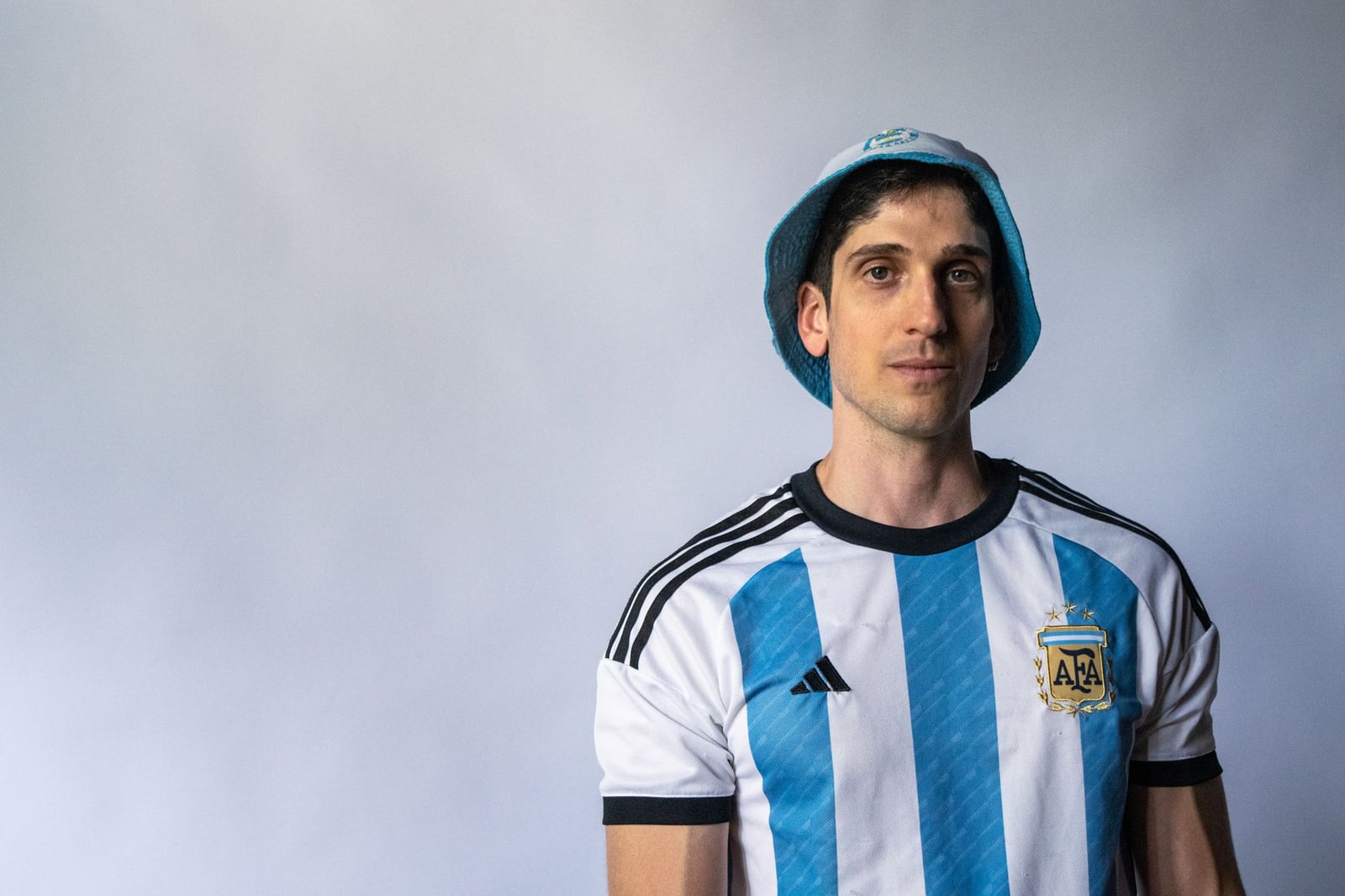 Retrato en primer plano de un hombre vestido con la camiseta y la gorra de fútbol argentino, sobre un fondo sencillo.