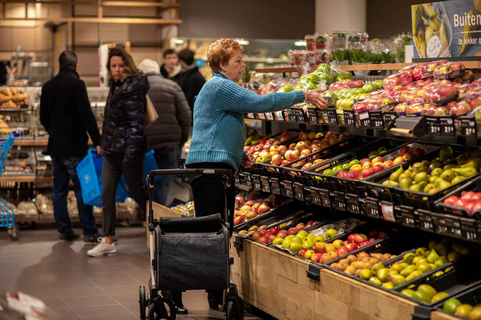 Mujer mayor usando andador, comprando productos frescos en un supermercado de Utrecht.