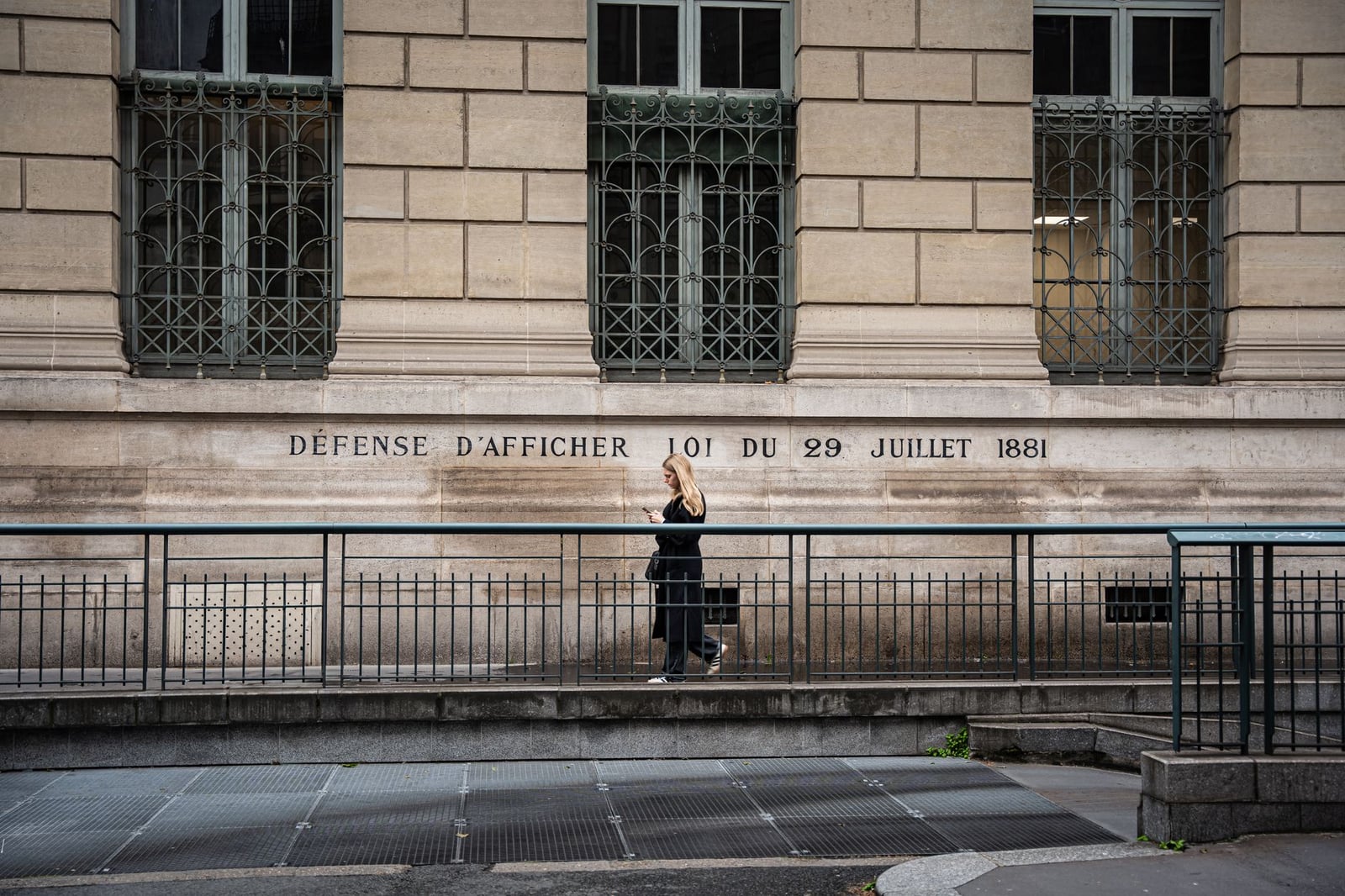 Mujer paseando por una calle de París junto a un edificio histórico con texto en francés.