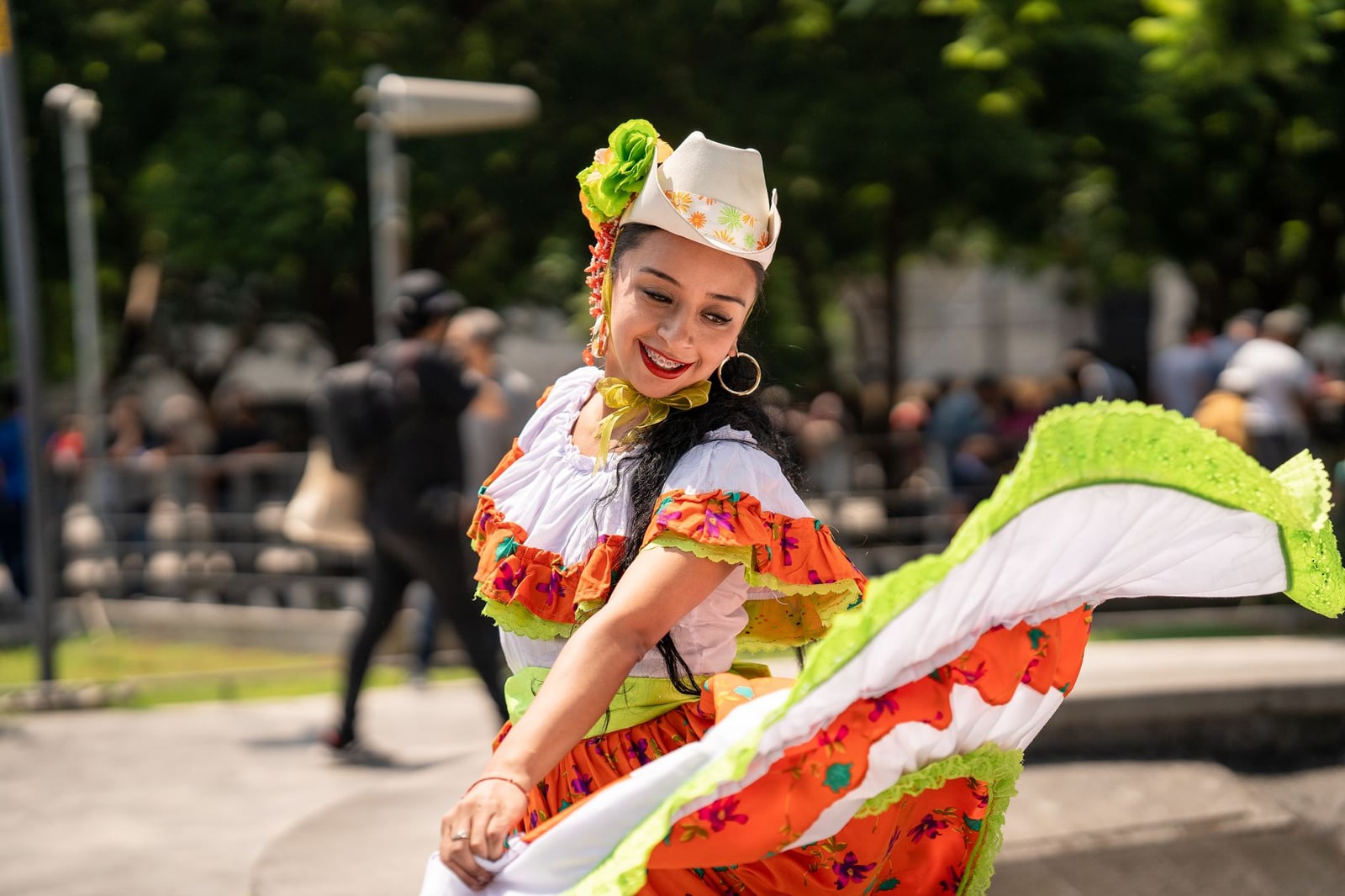Una mujer alegre con vestimenta tradicional colorida baila al aire libre durante un desfile de festival en un parque soleado.