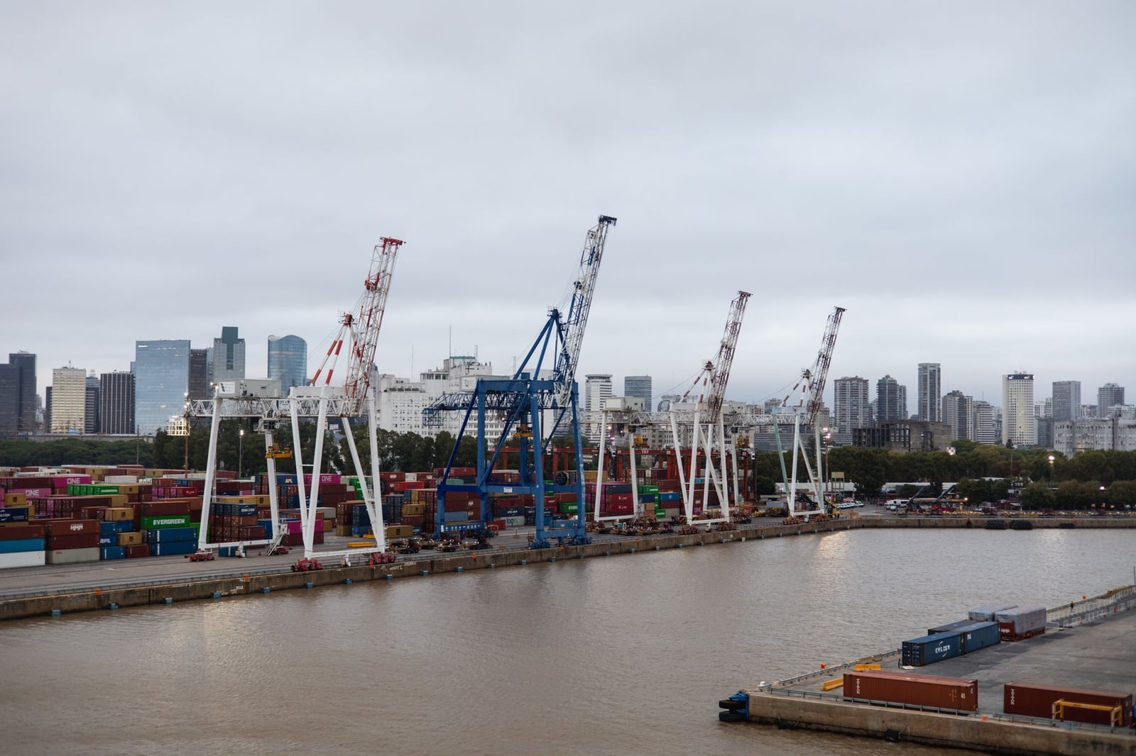 Contenedores de transporte y grúas en un puerto bullicioso con el perfil urbano de la ciudad de fondo.