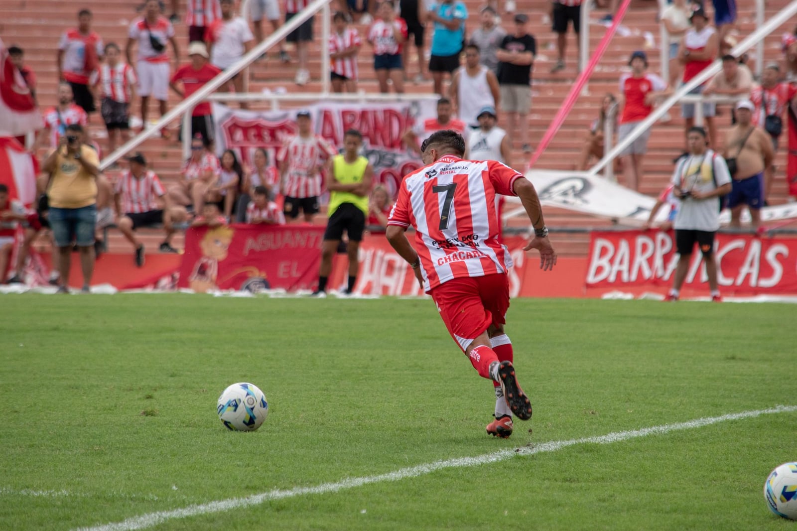 Un jugador de fútbol con rayas rojas y blancas juega durante un partido, con aficionados mirando desde las gradas.