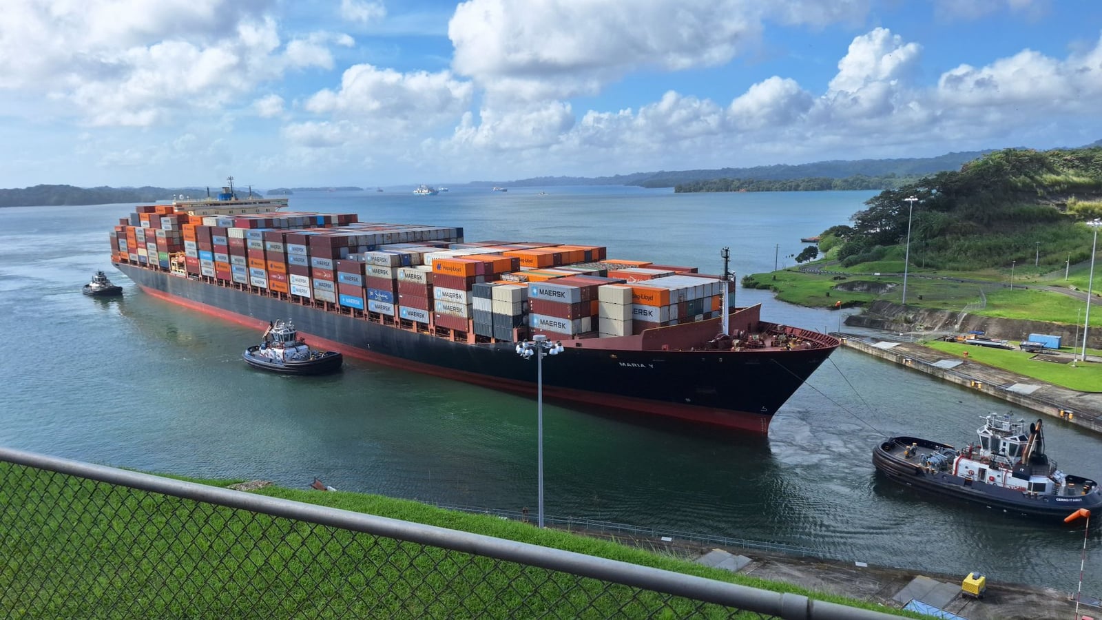 Gran buque portacontenedores maniobrado por remolcadores a través del Canal de Panamá bajo un cielo despejado.