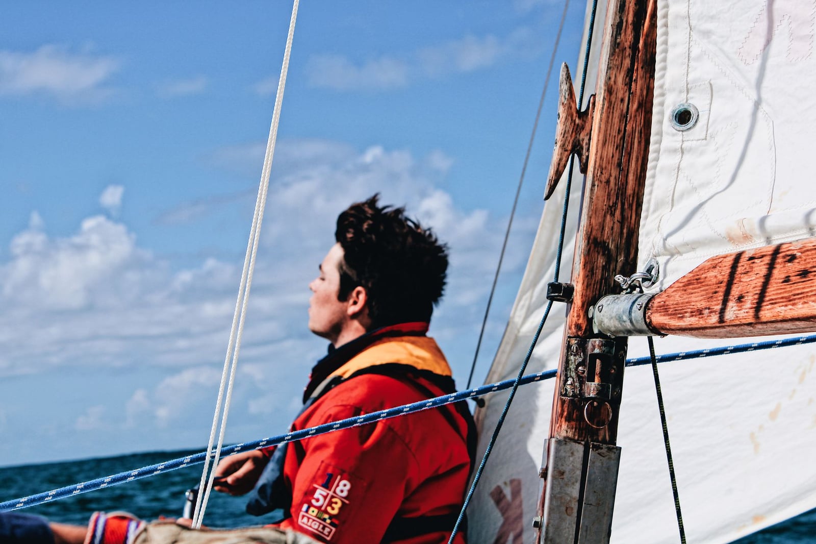 Un marinero solitario con una chaqueta roja mira al cielo mientras navega en un velero bajo cielos azules claros.