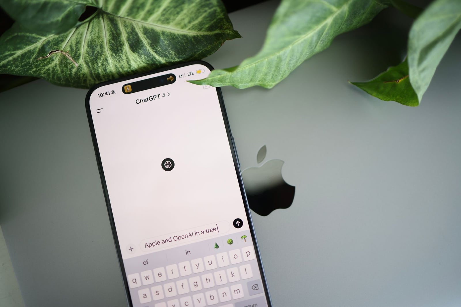 Un smartphone muestra una interfaz de ChatGPT colocada sobre un laptop Apple en un entorno con plantas.