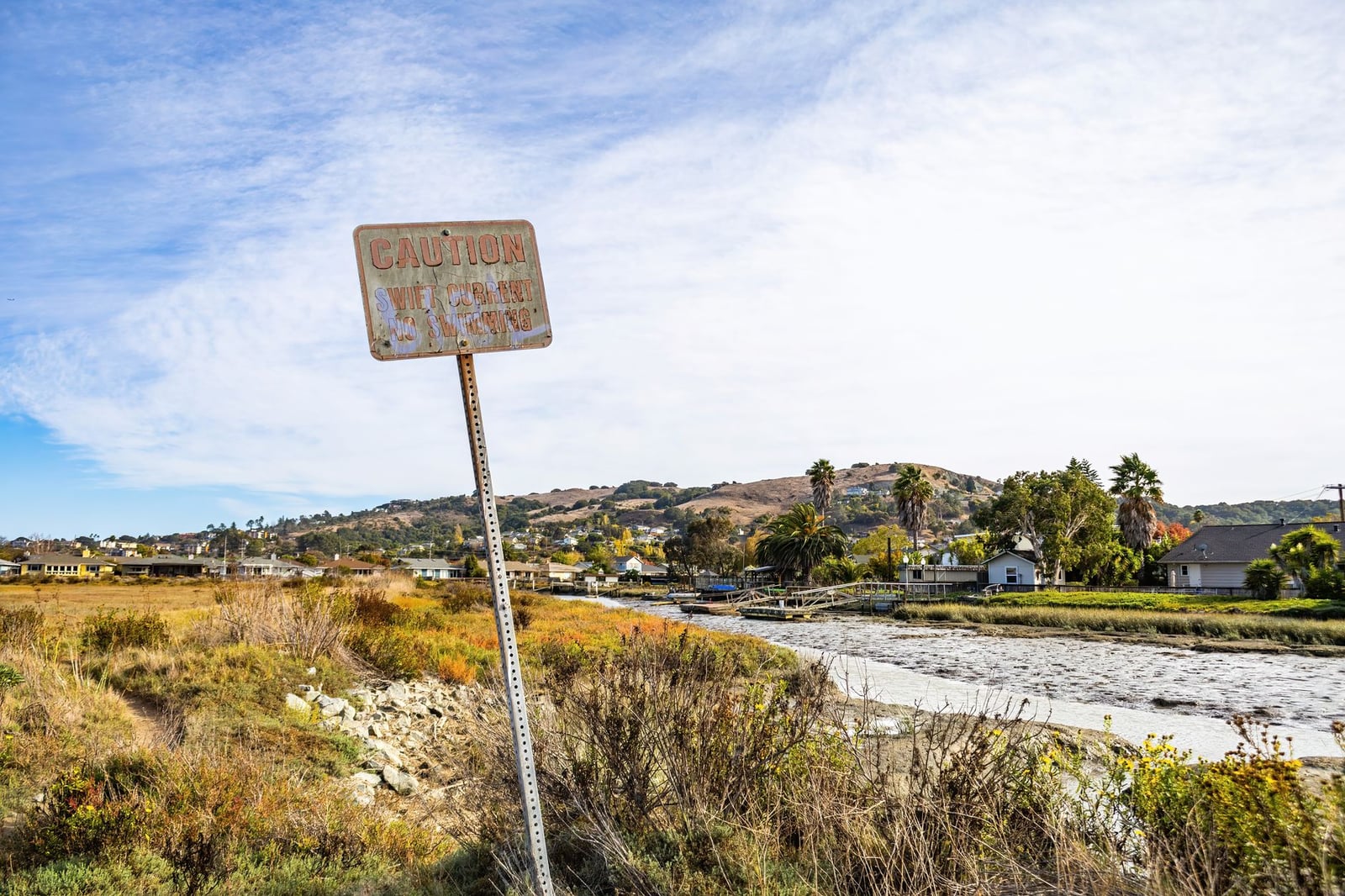 Escena rural con una señal de advertencia desgastada cerca de un río y casas.
