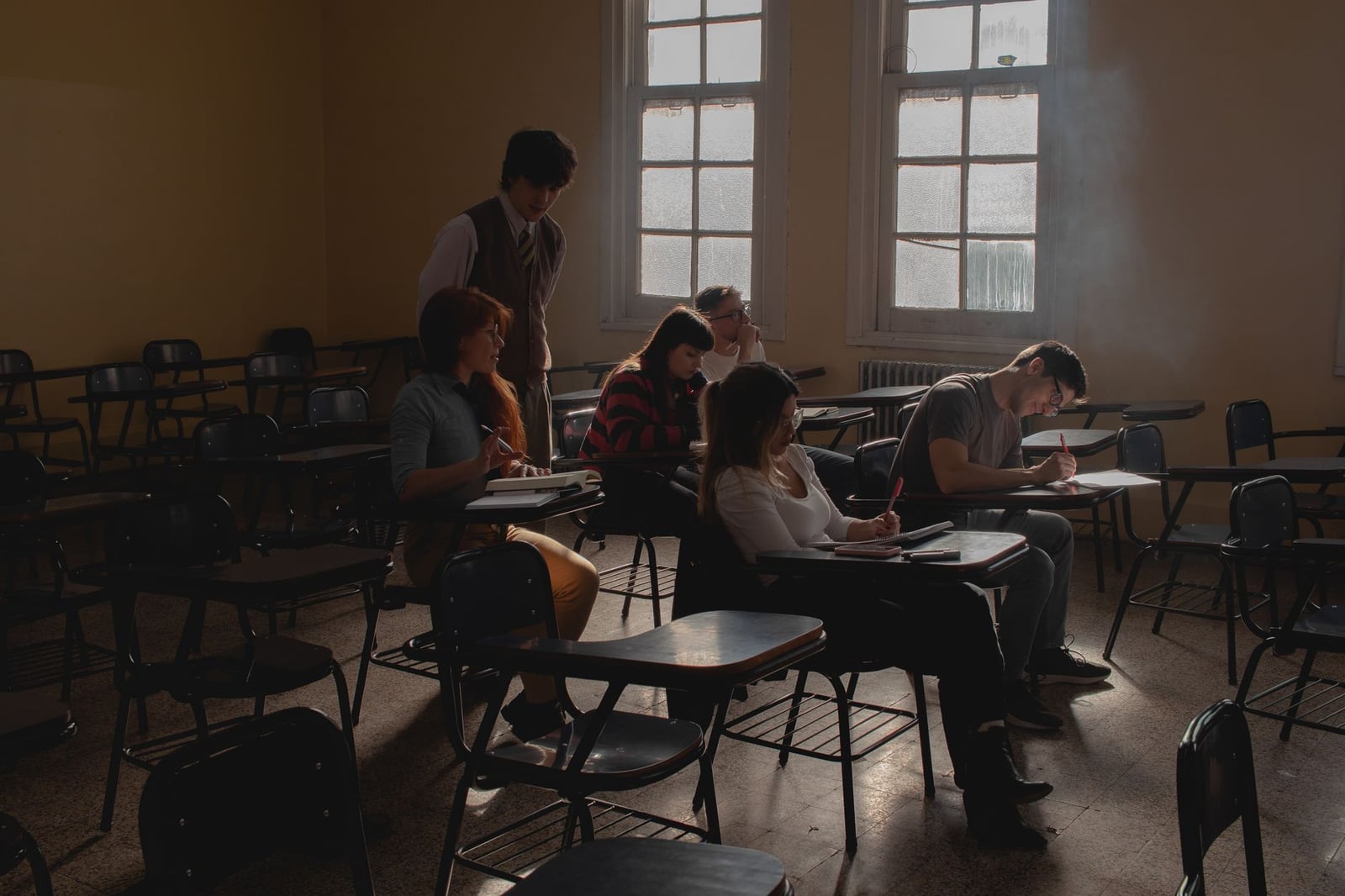 Grupo de estudiantes estudiando en un aula con poca luz, con pupitres antiguos y luz natural.