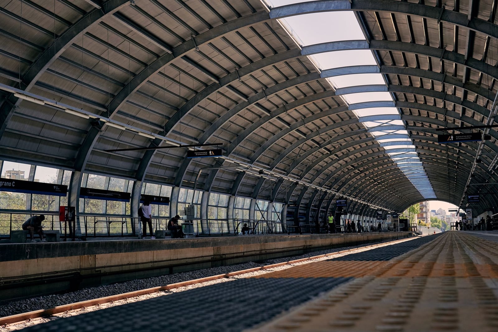 Plataforma moderna de estación de trenes con techo de acero curvado y pasajeros esperando.
