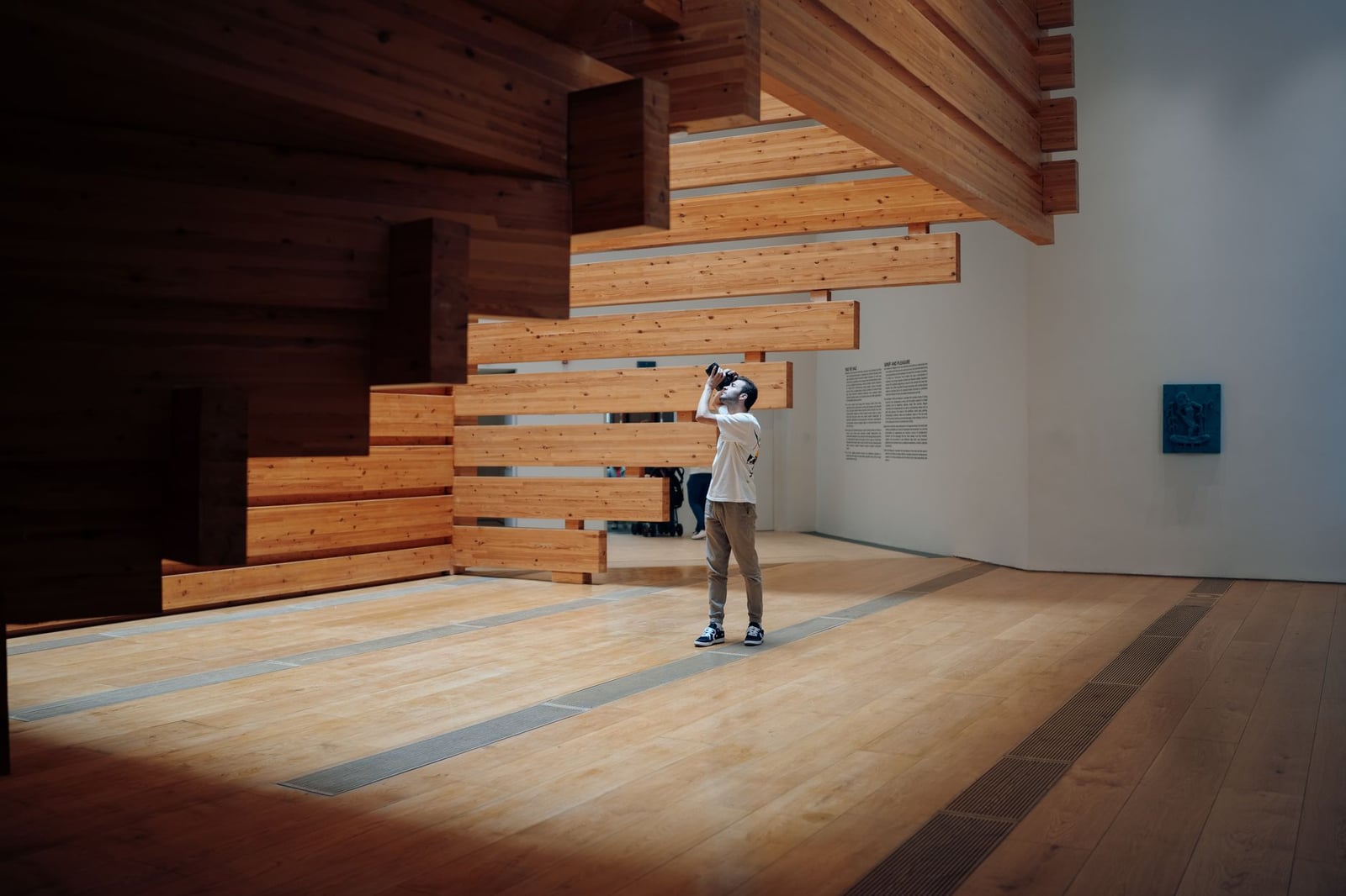 Un hombre fotografiando una instalación de arte en madera en un amplio entorno interior, destacando el diseño moderno.