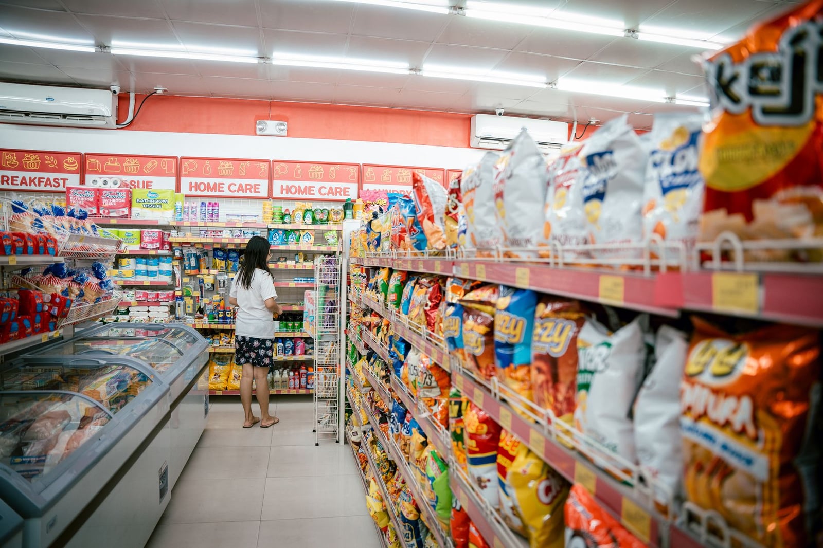Una mujer mirando las estanterías de snacks en un supermercado, rodeada de varios paquetes de patatas fritas.