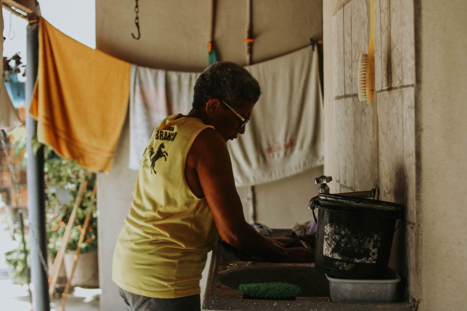 Mujer anciana con una camiseta amarilla lavando platos en un entorno doméstico.