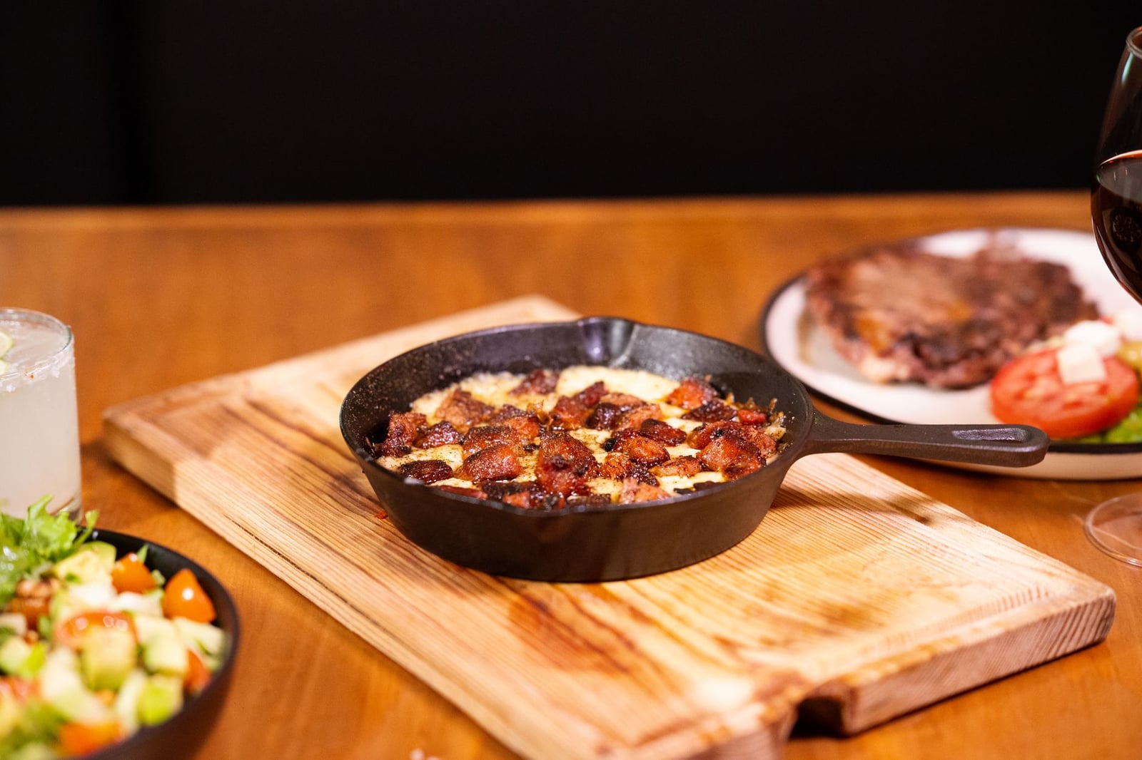 Plato exquisitamente preparado, servido en una sartén de hierro con ensalada y bebida, todo ello sobre una mesa de madera.