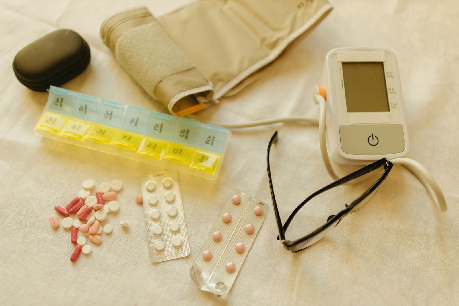 Imagen en plano cenital de elementos esenciales de salud, incluyendo un monitor de presión arterial, píldoras y gafas sobre una superficie blanca.