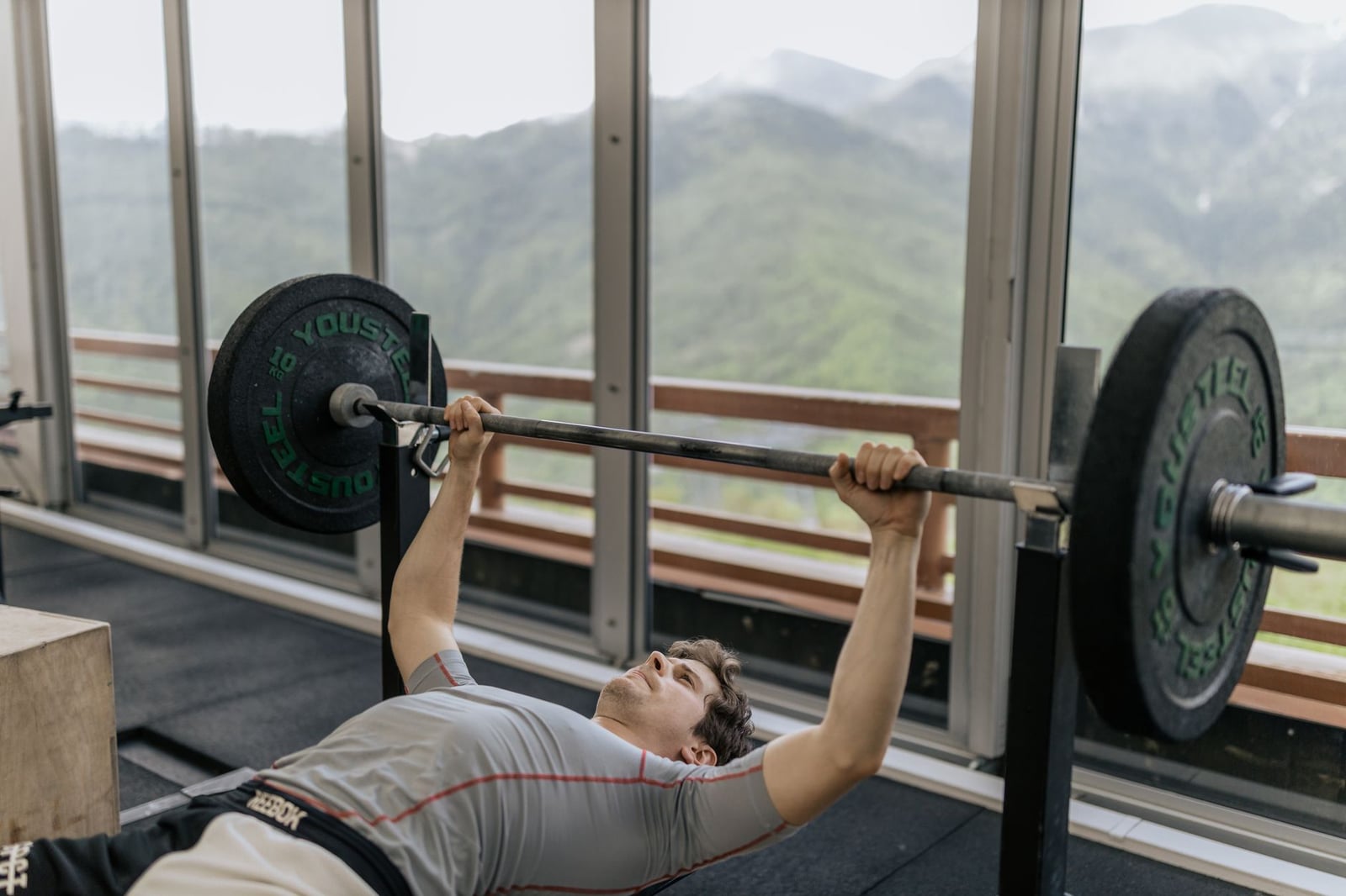 Un hombre realiza un press de banca en un gimnasio con vistas a montañas, enfatizando la fuerza y la acondicionamiento físico.