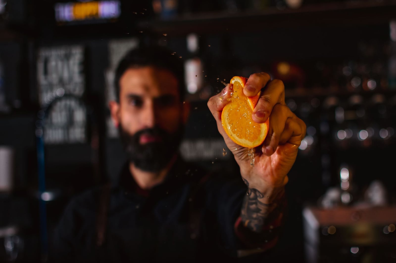 Barman con barba exprime una cuña de naranja, mostrando un jugo vibrante en un bar con poca luz.
