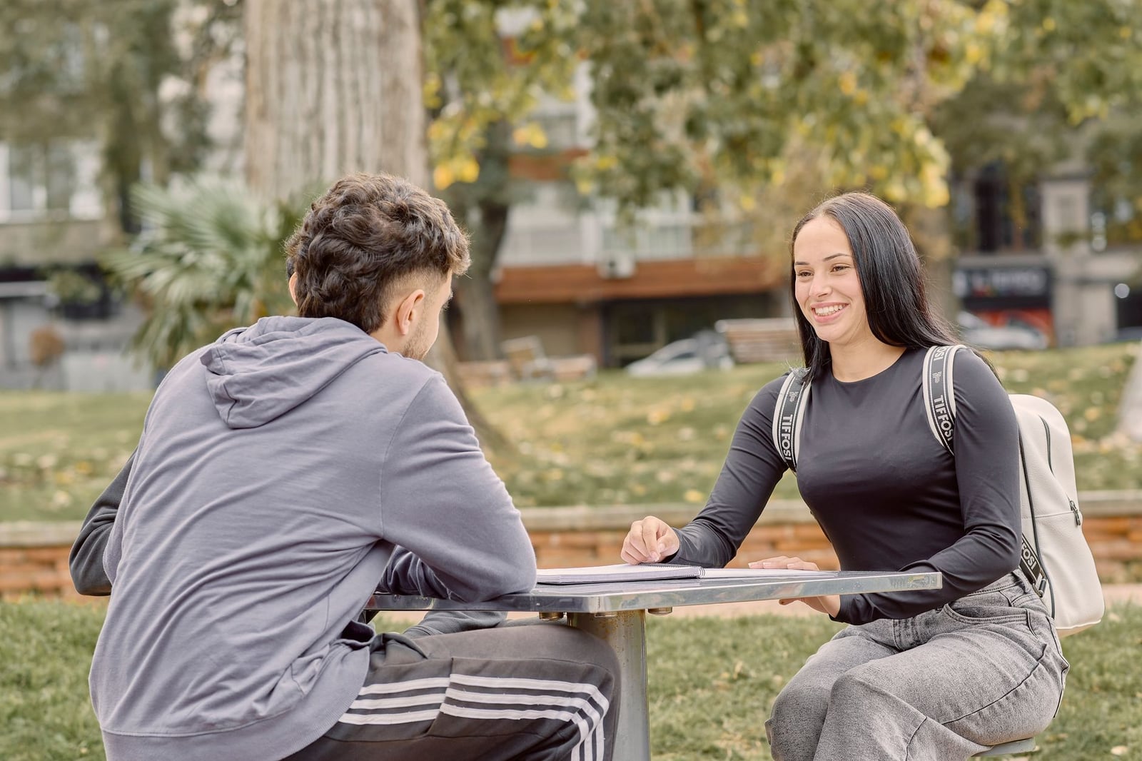 Dos jóvenes adultos sentados en una mesa al aire libre, sonriendo y conversando en un entorno de parque.