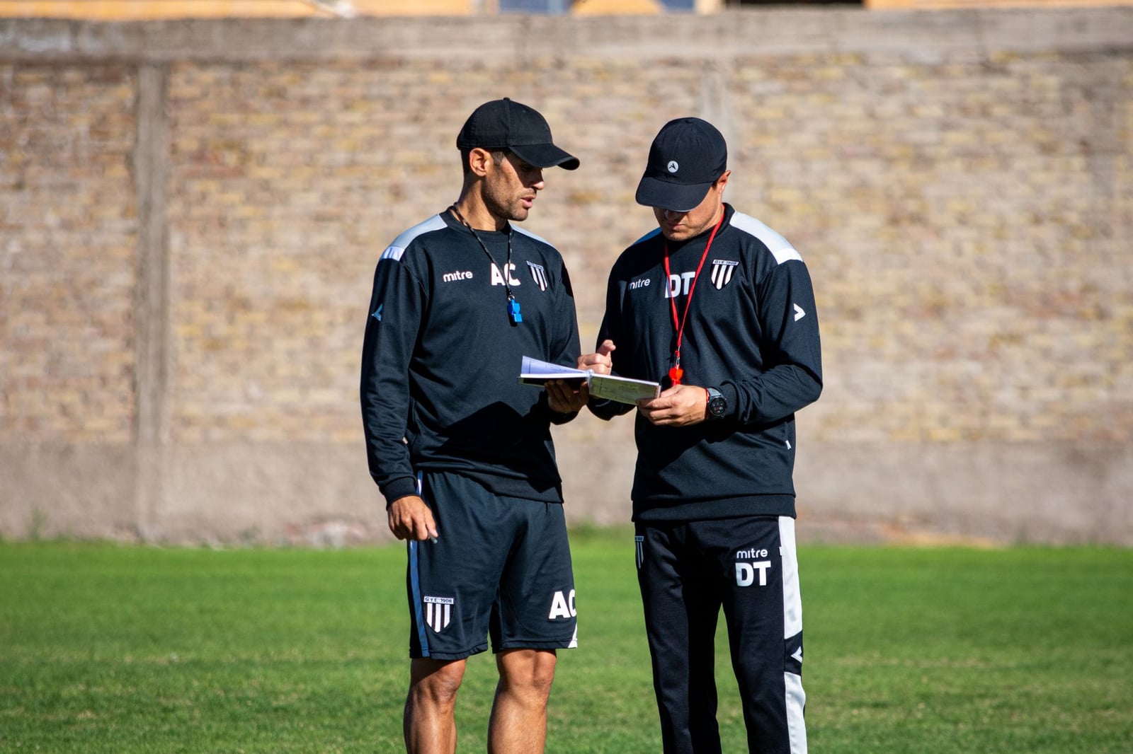 Dos entrenadores de fútbol revisando estrategias en el campo durante el día.