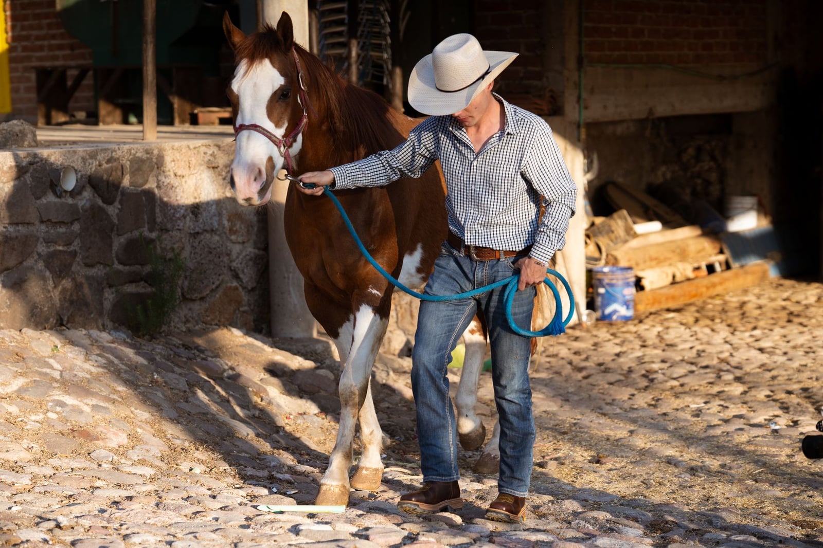 Un vaquero con sombrero guía un caballo castaño por un camino empedrado a las afueras de un establo.
