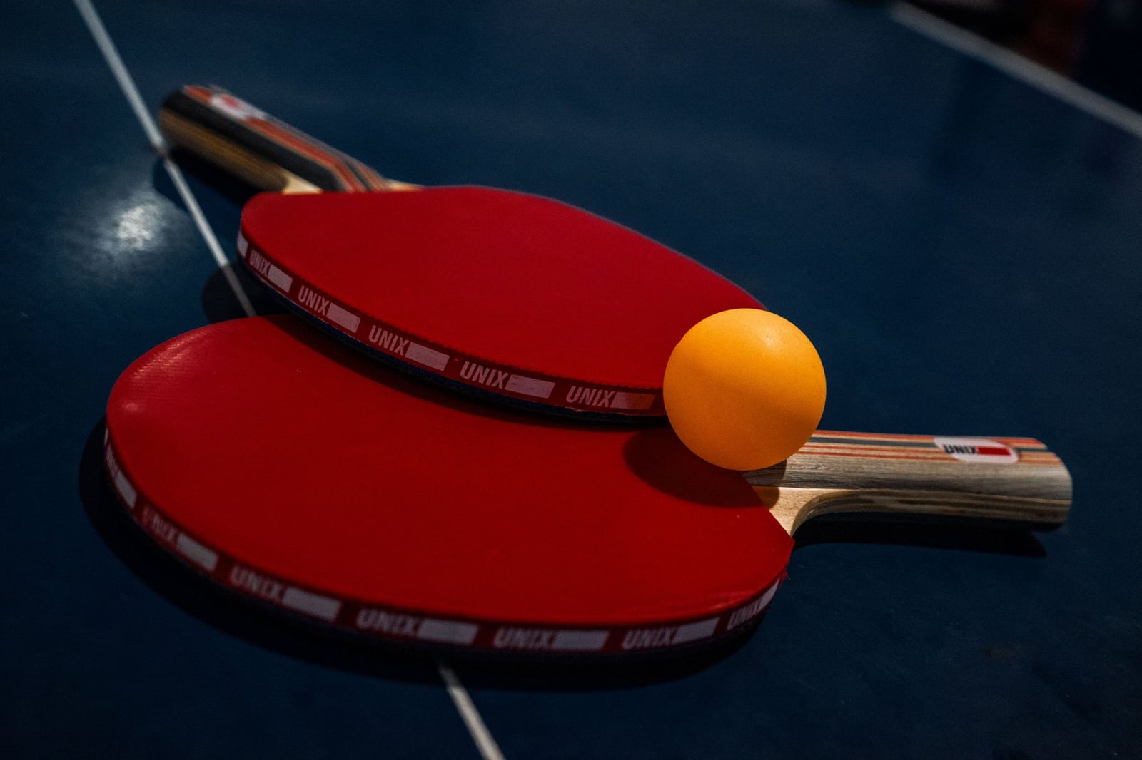 Primer plano de dos palas de ping-pong rojas y una pelota amarilla sobre una mesa azul oscuro.