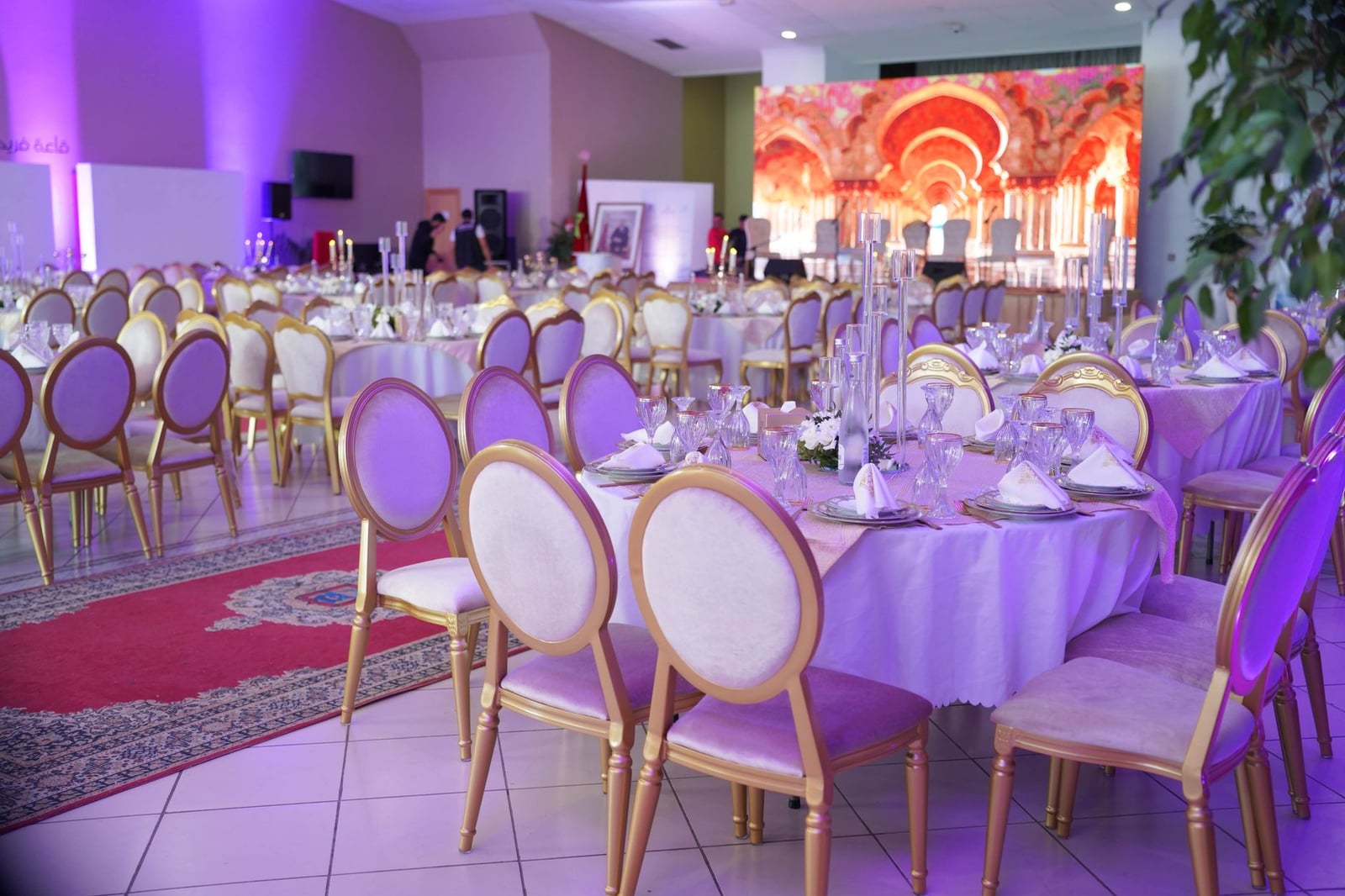Sala de banquete lujosa preparada para un gran evento con decoración elegante, iluminación púrpura y mesas dispuestas.