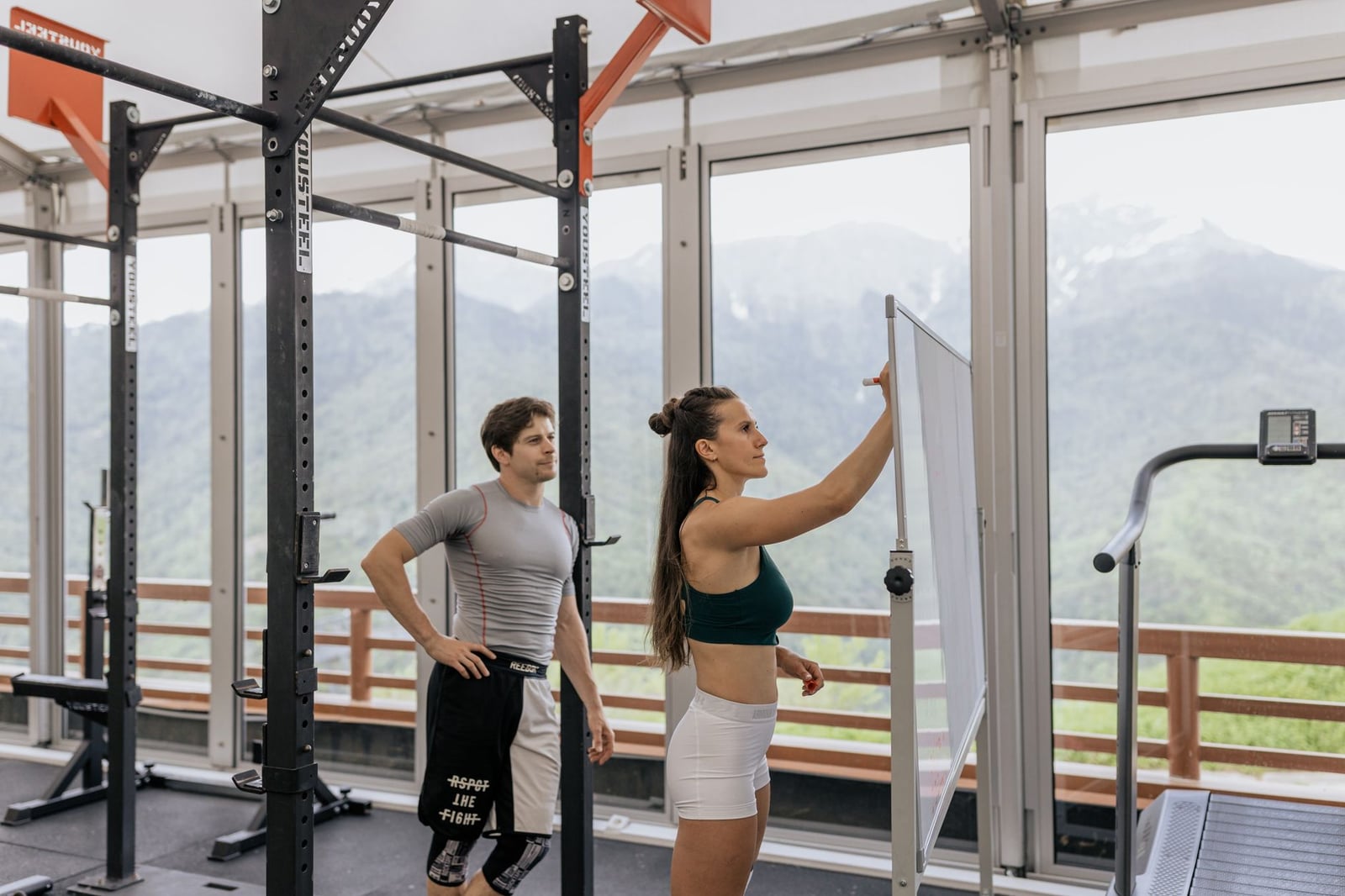 Un entrenador de gimnasio instruye en una pizarra junto a equipos de fitness con vista a las montañas.