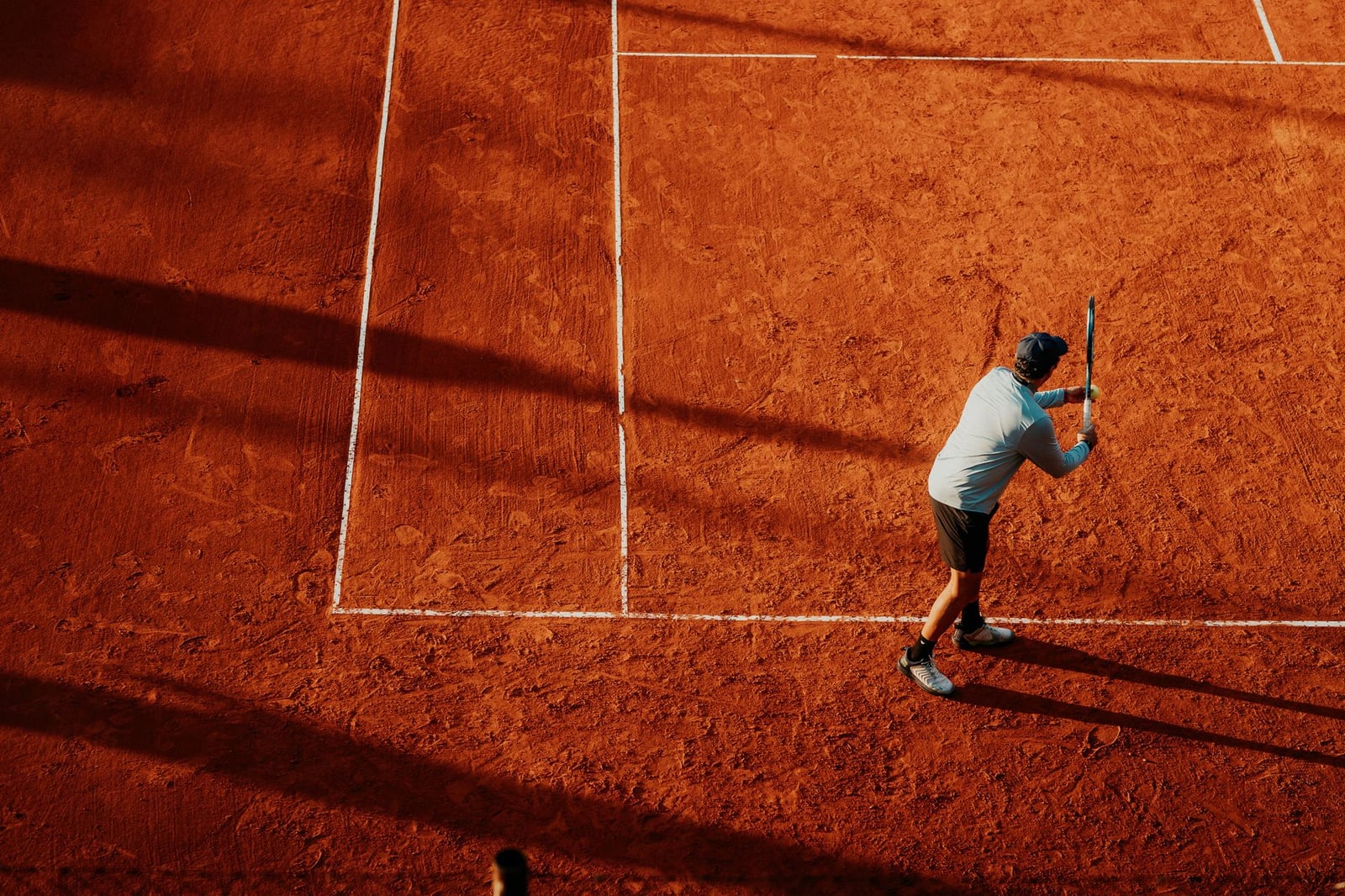 Vista aérea de un jugador de tenis en una cancha de arcilla en Buenos Aires, proyectando largas sombras.