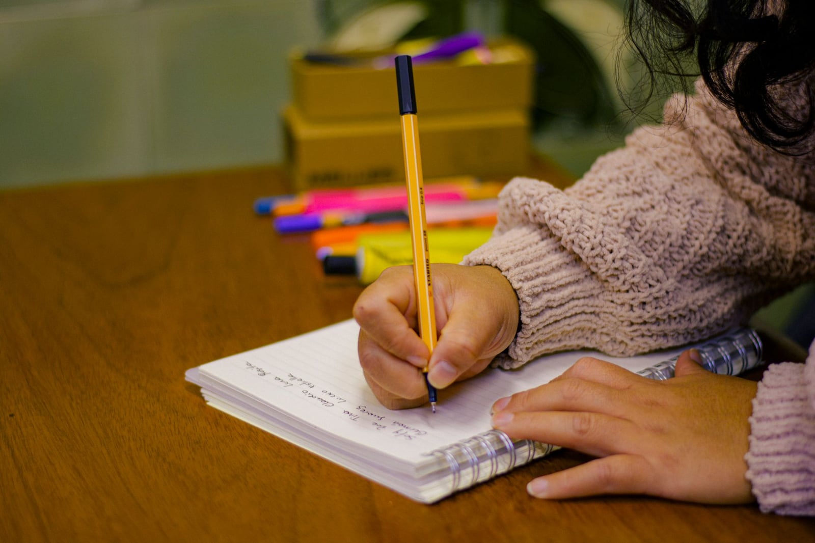 Una persona escribiendo en un cuaderno con bolígrafos y resaltadores en un escritorio de madera, creando una escena de estudio.