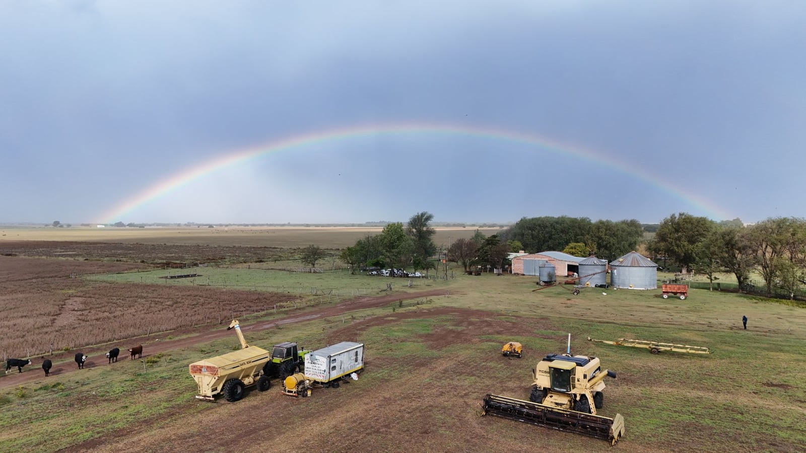 Un arcoíris vibrante se arquea sobre una granja rural con maquinaria y graneros en La Pampa, creando una pintoresca escena agrícola.