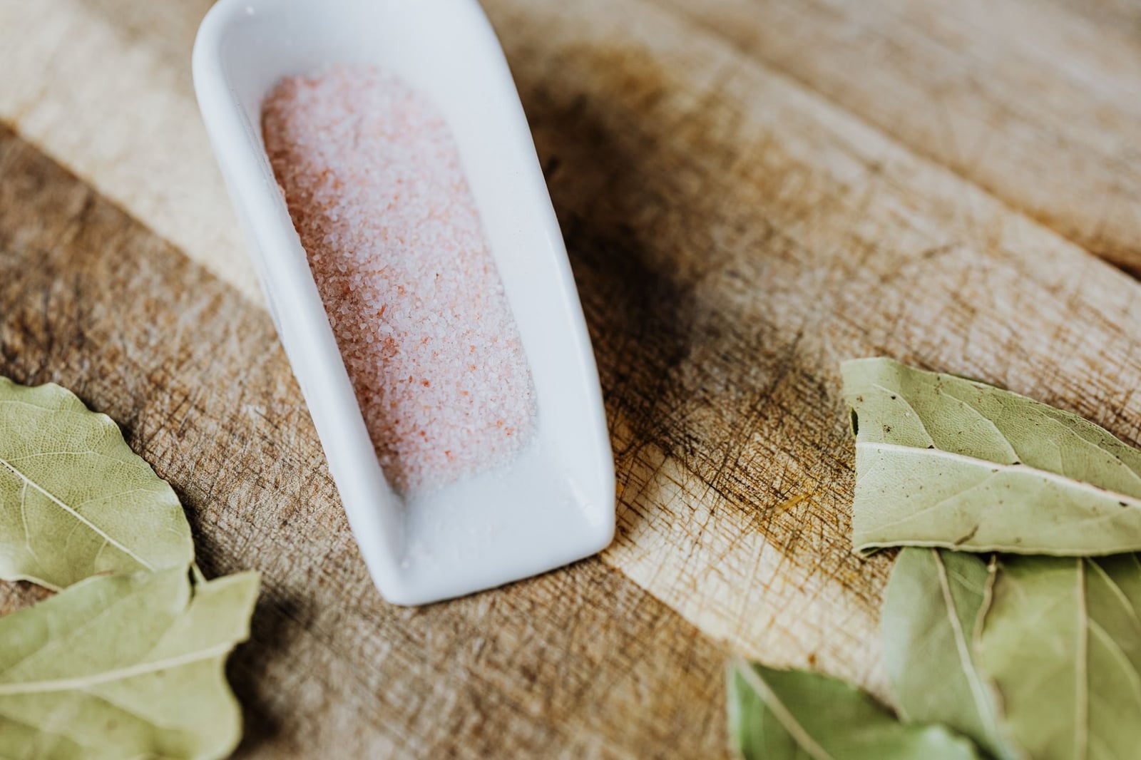 Primer plano de sal rosa del Himalaya y hojas de laurel sobre una superficie de madera rústica, destacando ingredientes naturales.