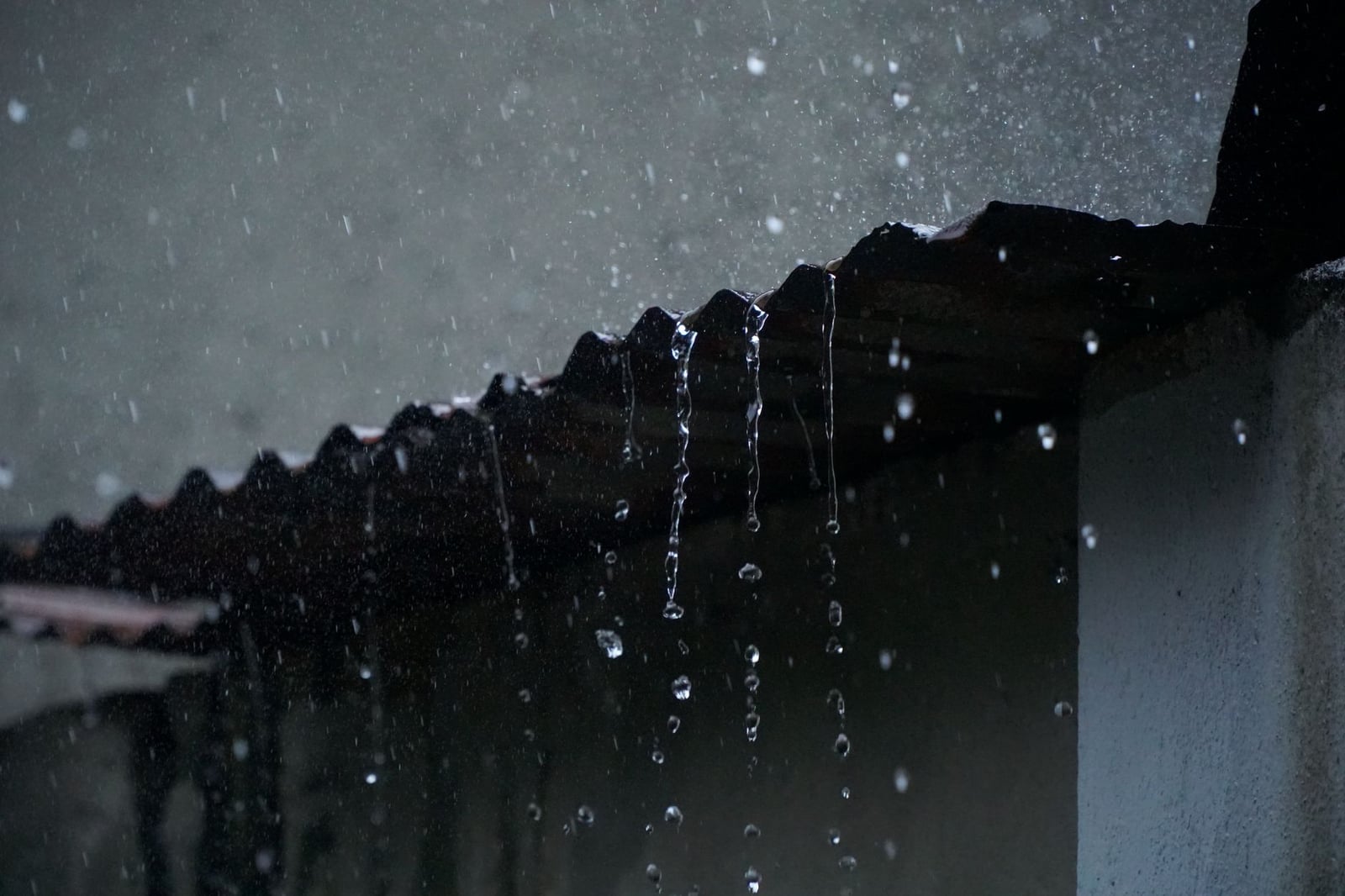 Primer plano del agua de lluvia fluyendo de un tejado corrugado en poca luz, creando una atmósfera serena.