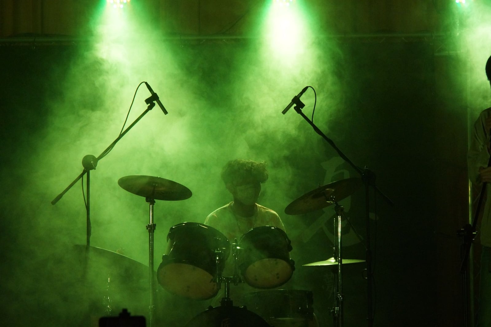 Un baterista actúa en el escenario entre humo y luces verdes, creando una atmósfera vibrante de concierto.