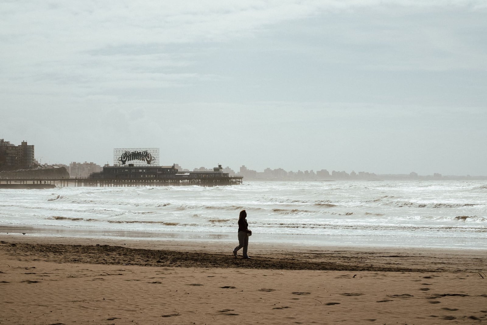 Una persona solitaria camina por una playa desierta con un muelle antiguo al fondo.