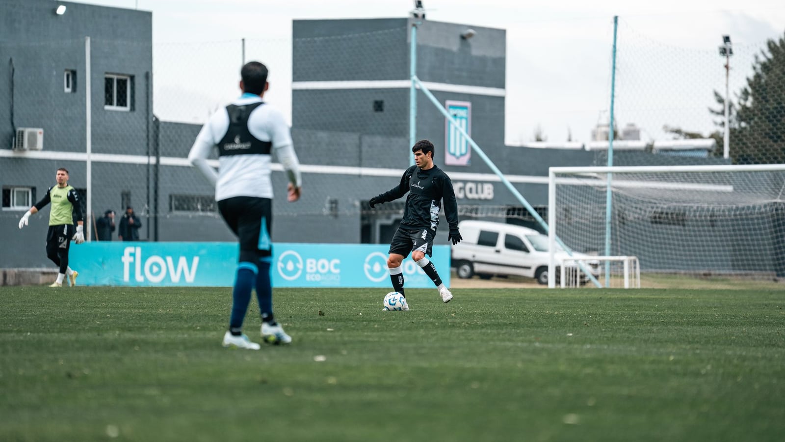 Dos jugadores de fútbol practicando en un campo durante una sesión de entrenamiento, con césped verde exuberante y un poste de gol visible.