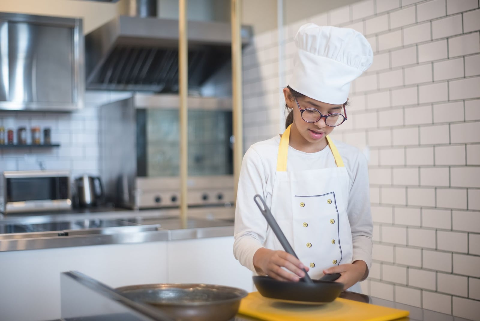 Una niña joven vestida con atuendo de chef cocinando en una cocina moderna con una sartén.