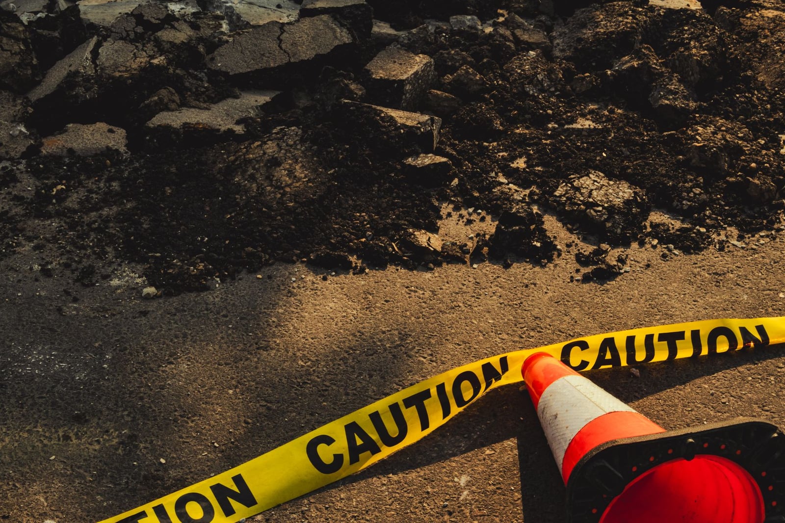 Acera rota con escombros, cinta de precaución y cono de tráfico en sitio de construcción urbana.