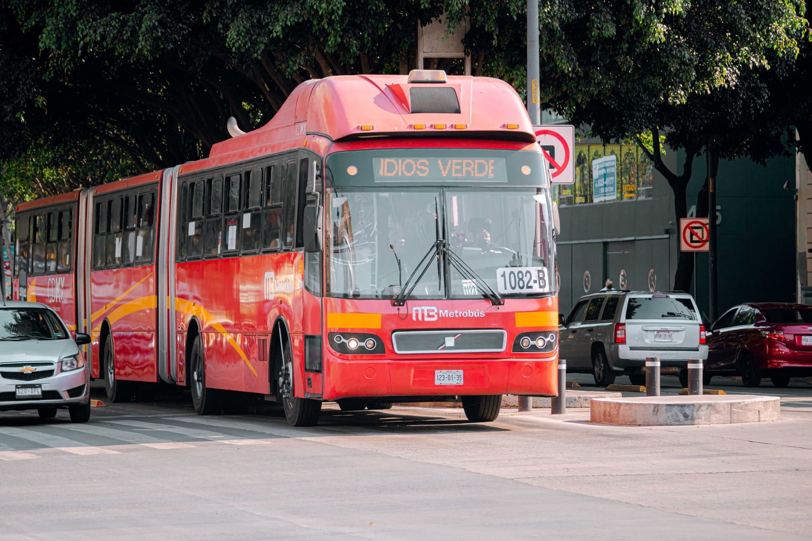 Un Metrobús rojo navegando una calle de la ciudad rodeado de coches y vegetación.