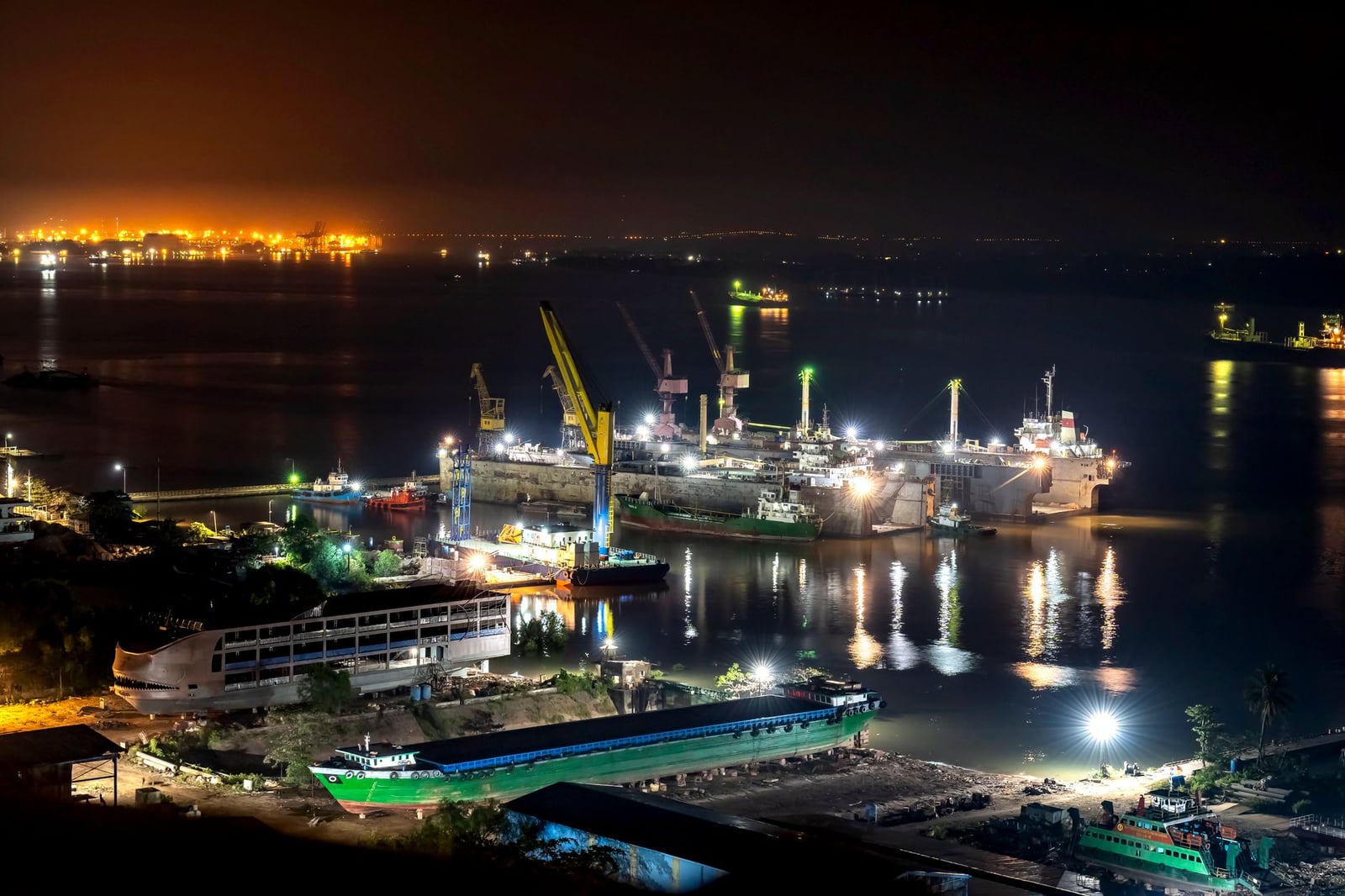 Una vibrante vista nocturna de un puerto industrial ocupado con grúas iluminadas y reflejos en el agua.