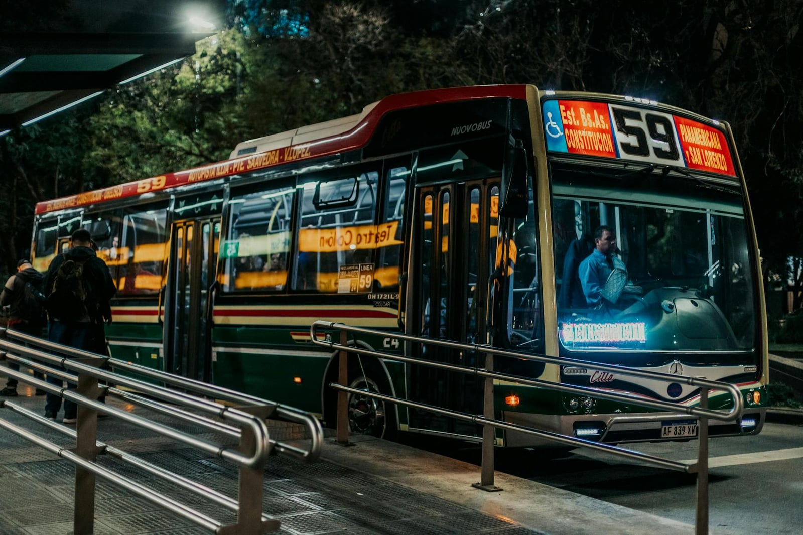 Una escena de una parada de autobús urbano por la noche con un autobús estacionado y pasajeros subiendo.