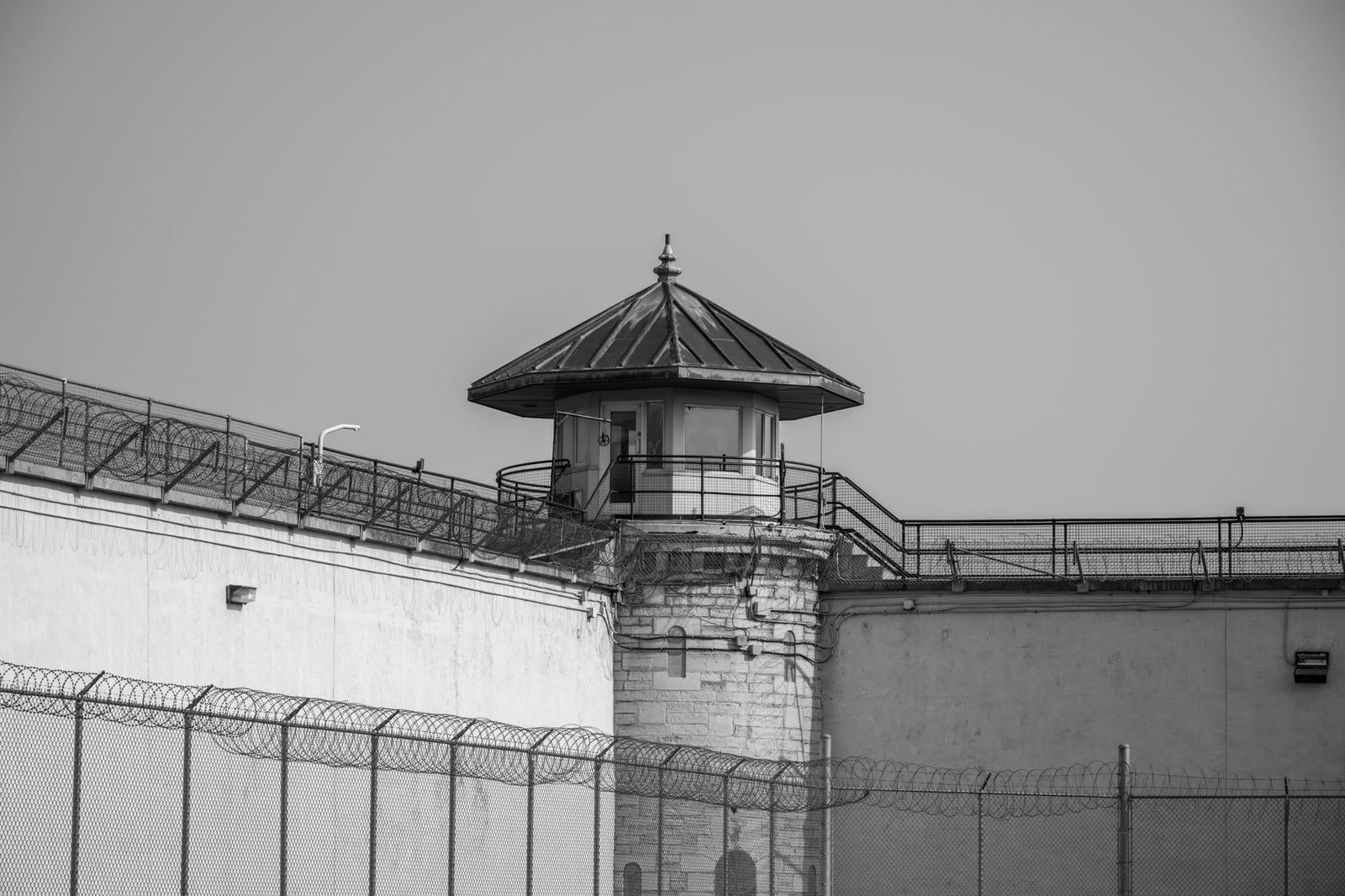 Imagen en blanco y negro de una torre de vigilancia de prisión con cercas de alambre de espino y medidas de seguridad bajo un cielo despejado.
