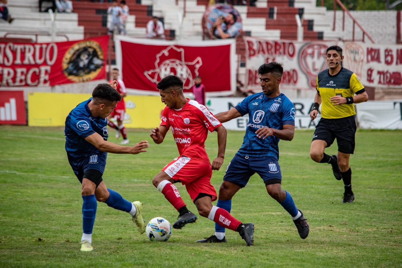 Jugadores de fútbol en acción durante un partido competitivo en un campo de hierba con espectadores en las gradas.