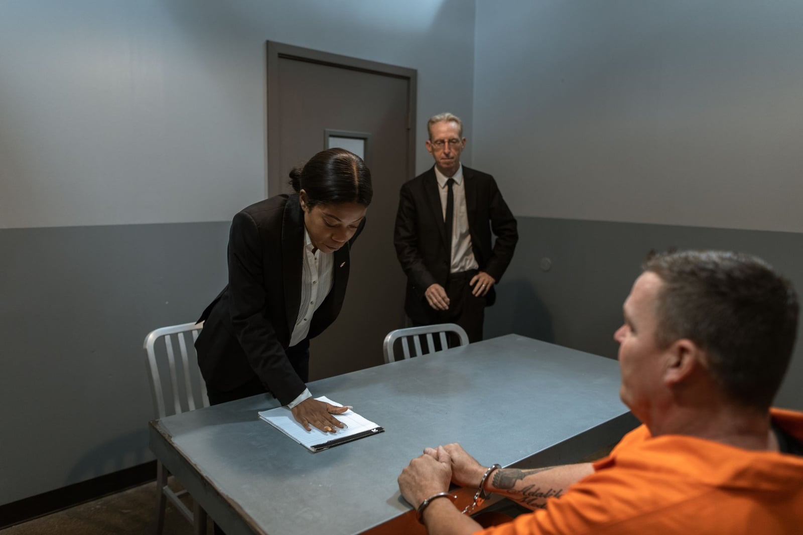 Un prisionero con esposas es interrogado por un detective y un abogado en una sala de interrogatorio.
