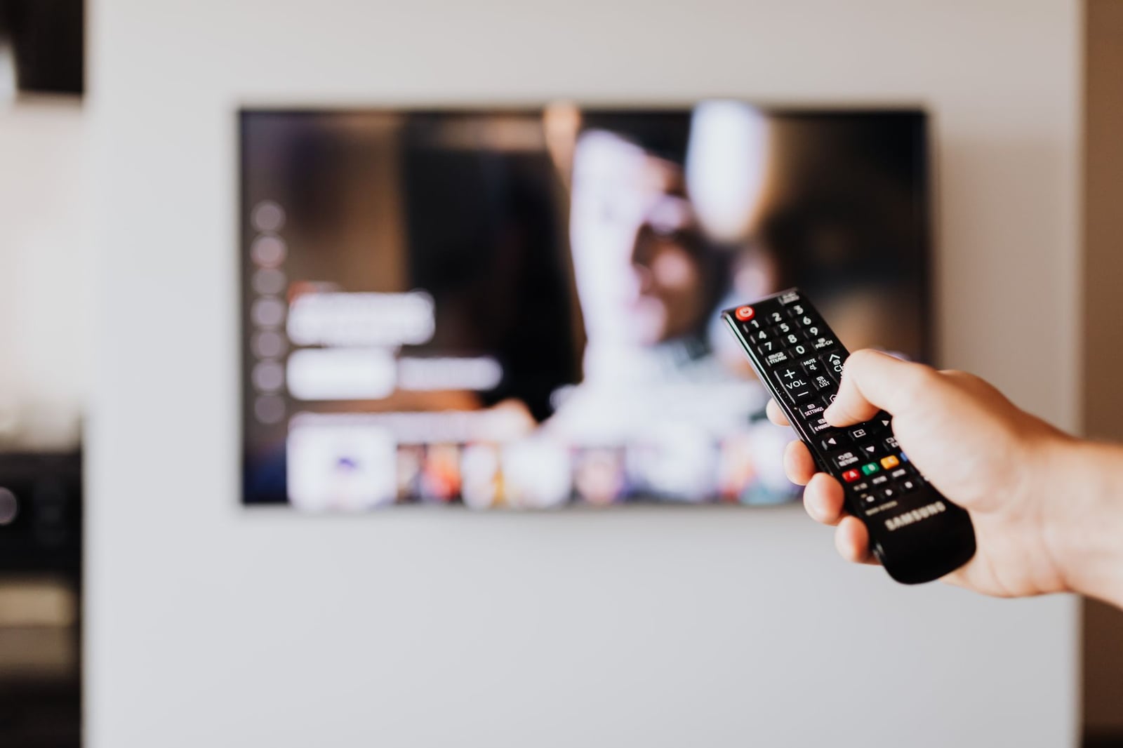 Persona usando un control remoto de televisión con una pantalla de televisión desenfocada al fondo.