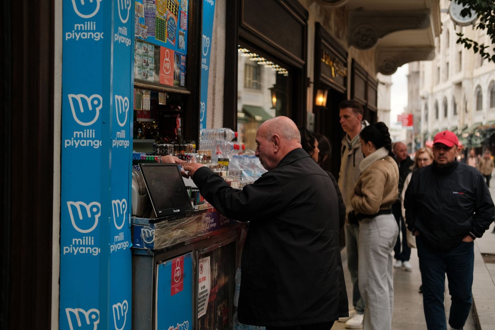 Personas comprando billetes de lotería y bebidas a un vendedor ambulante en una bulliciosa calle urbana.