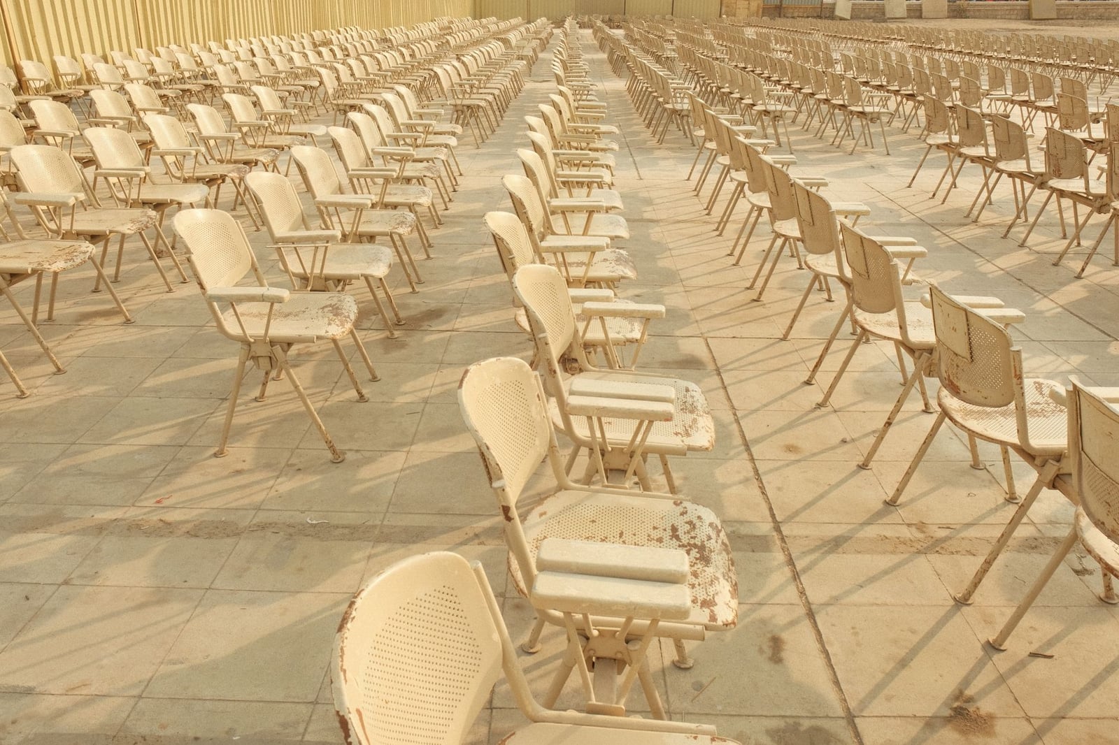 Escenario al aire libre con filas de sillas blancas desgastadas bajo la luz del sol.