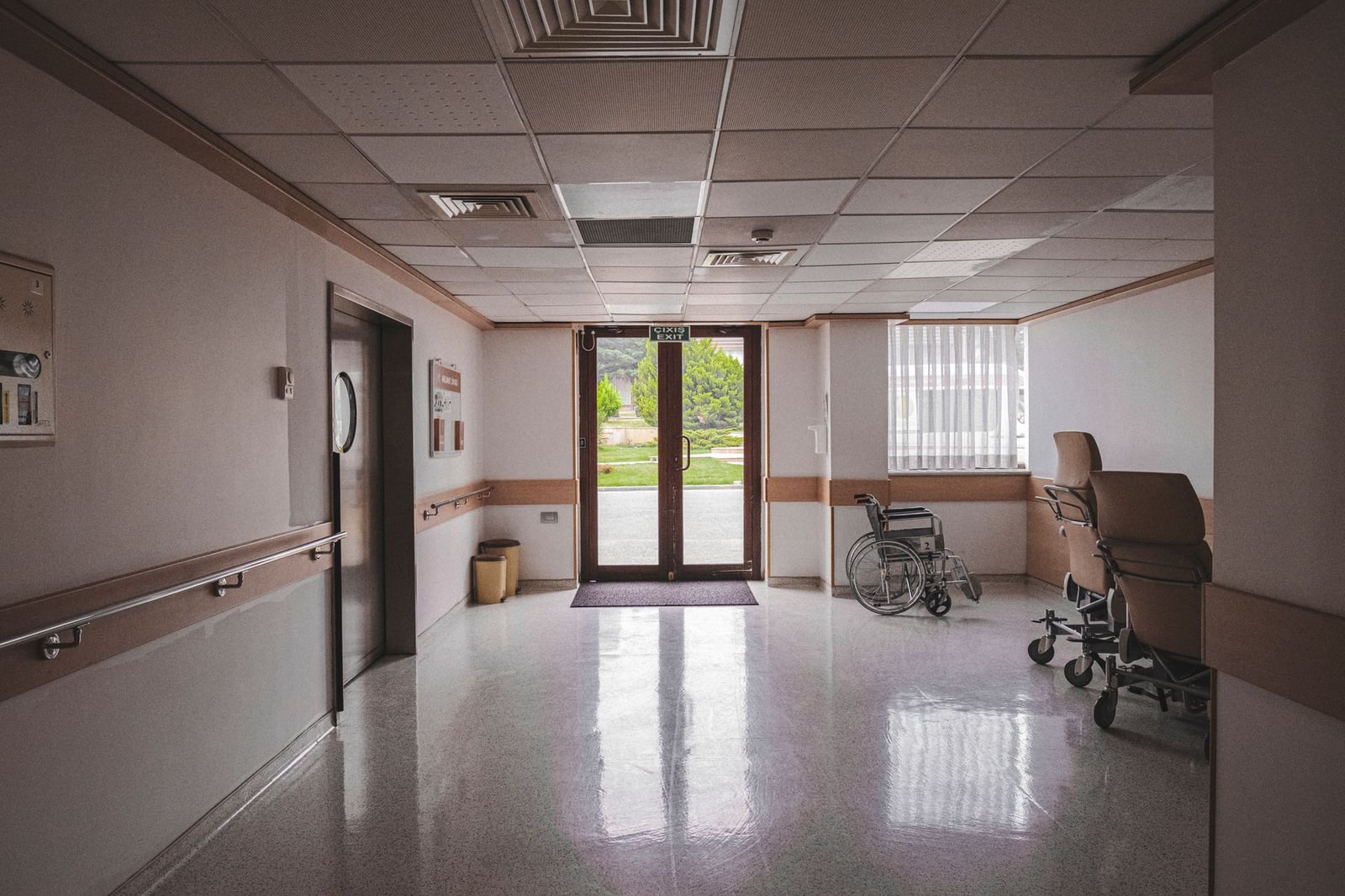 Pasillo de hospital con silla de ruedas, puertas de cristal reflejantes y vista exterior. Ideal para temas de atención médica.