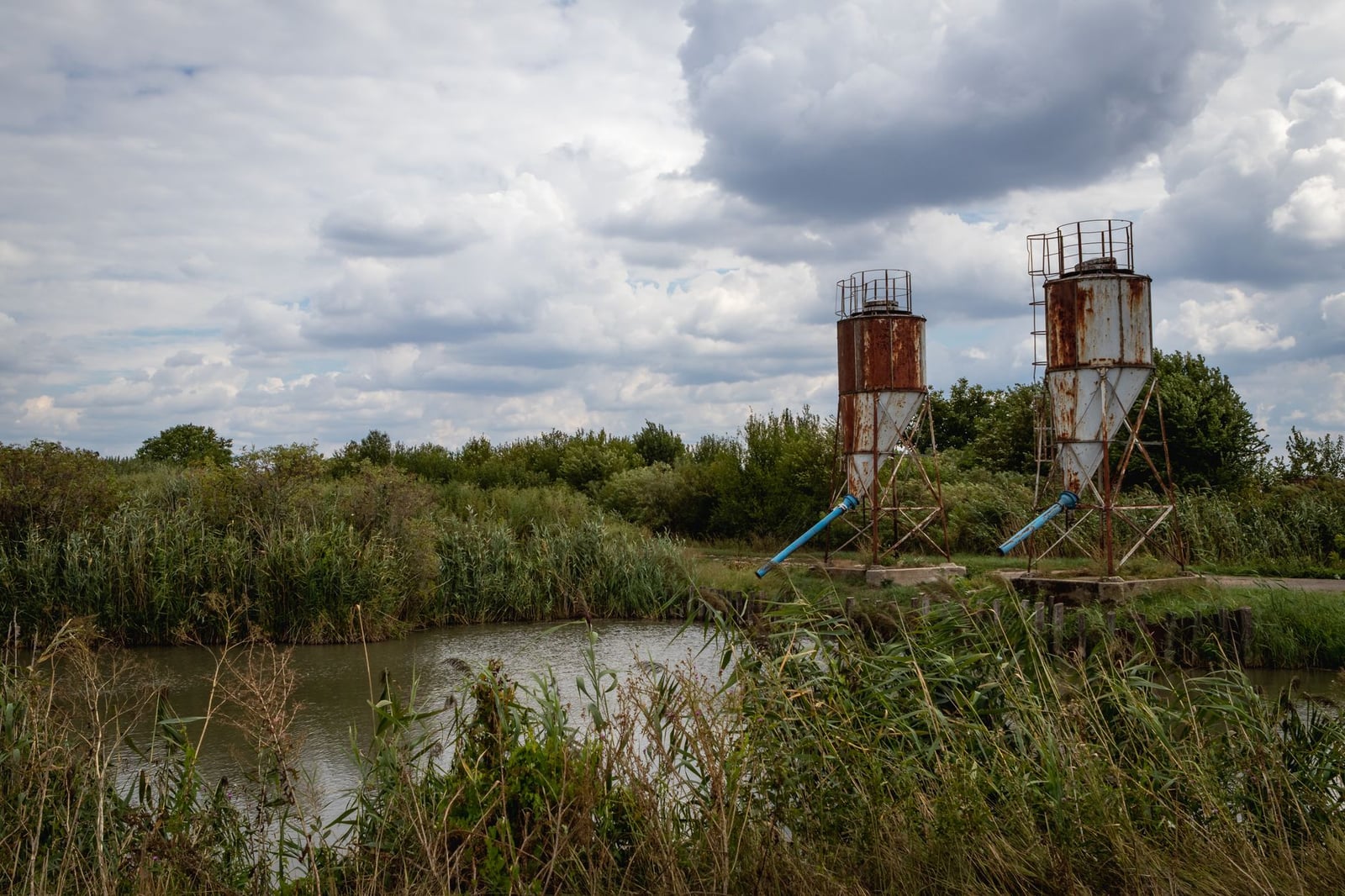 Dos silos oxidados se encuentran junto a un pantano bajo un cielo nublado en una zona rural.