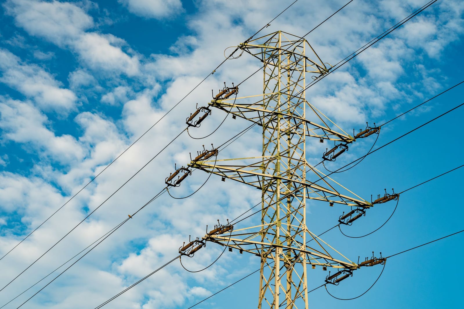 Una alta torre de transmisión eléctrica se alza sobre un fondo de cielo azul y nubes blancas.