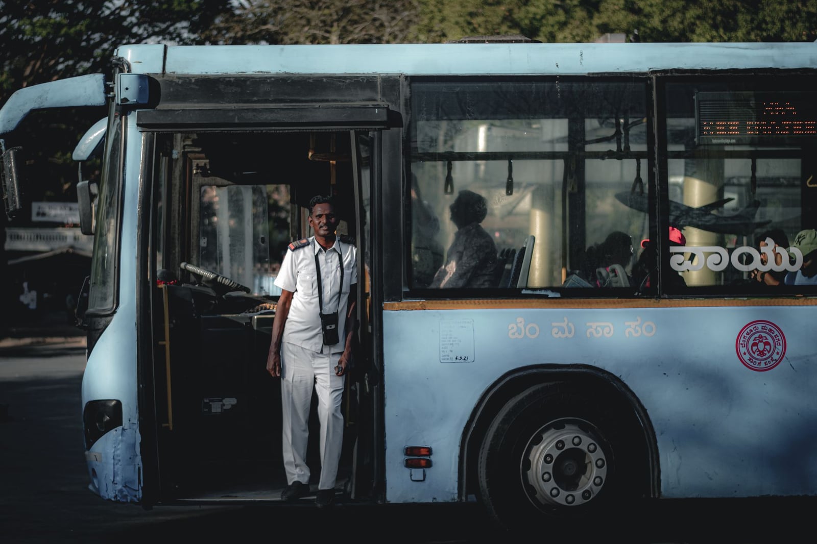 Conductor de autobús con uniforme blanco de pie junto a la puerta del autobús, la luz del sol proyectando sombras en el interior.