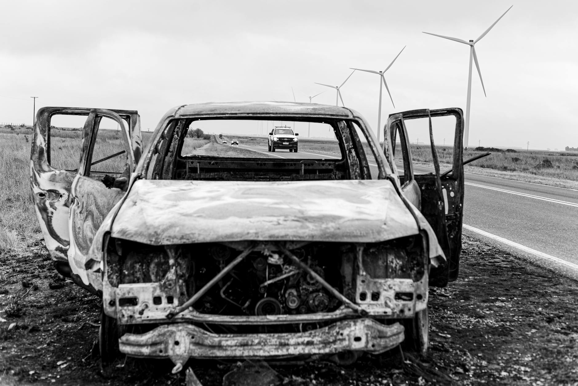 Imagen monocromática de un automóvil quemado al costado de la carretera con turbinas eólicas en Buenos Aires.